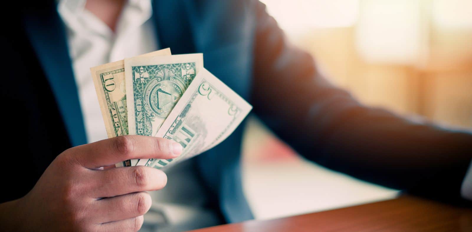A cheerful woman in a teal blazer laughs while holding a large fan of $100 bills near a laptop in a bright office