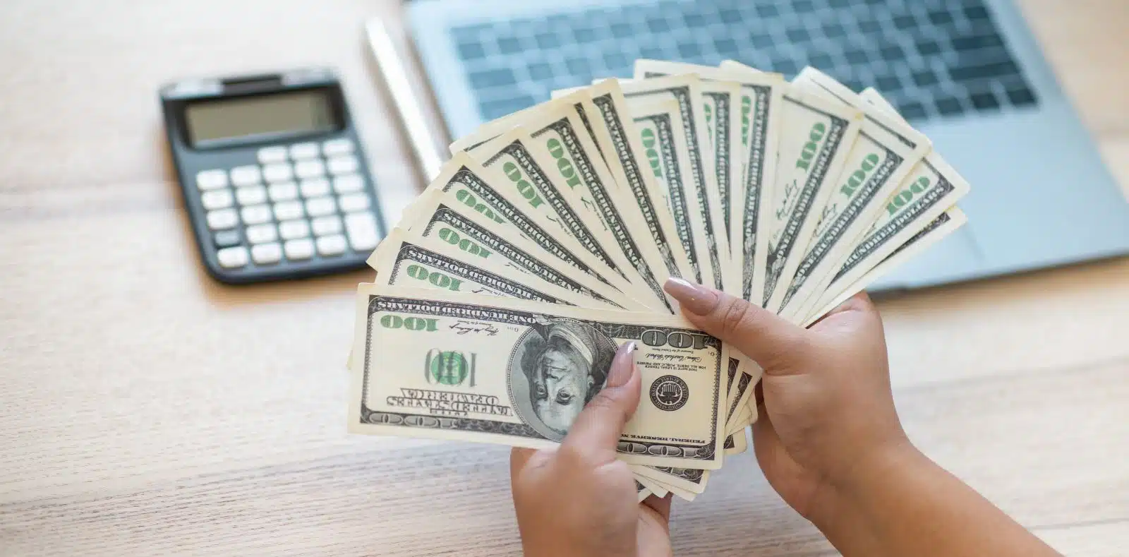 A hand offers a folded stack of $100 bills to a smiling young woman in a pink cardigan against a bright pink background