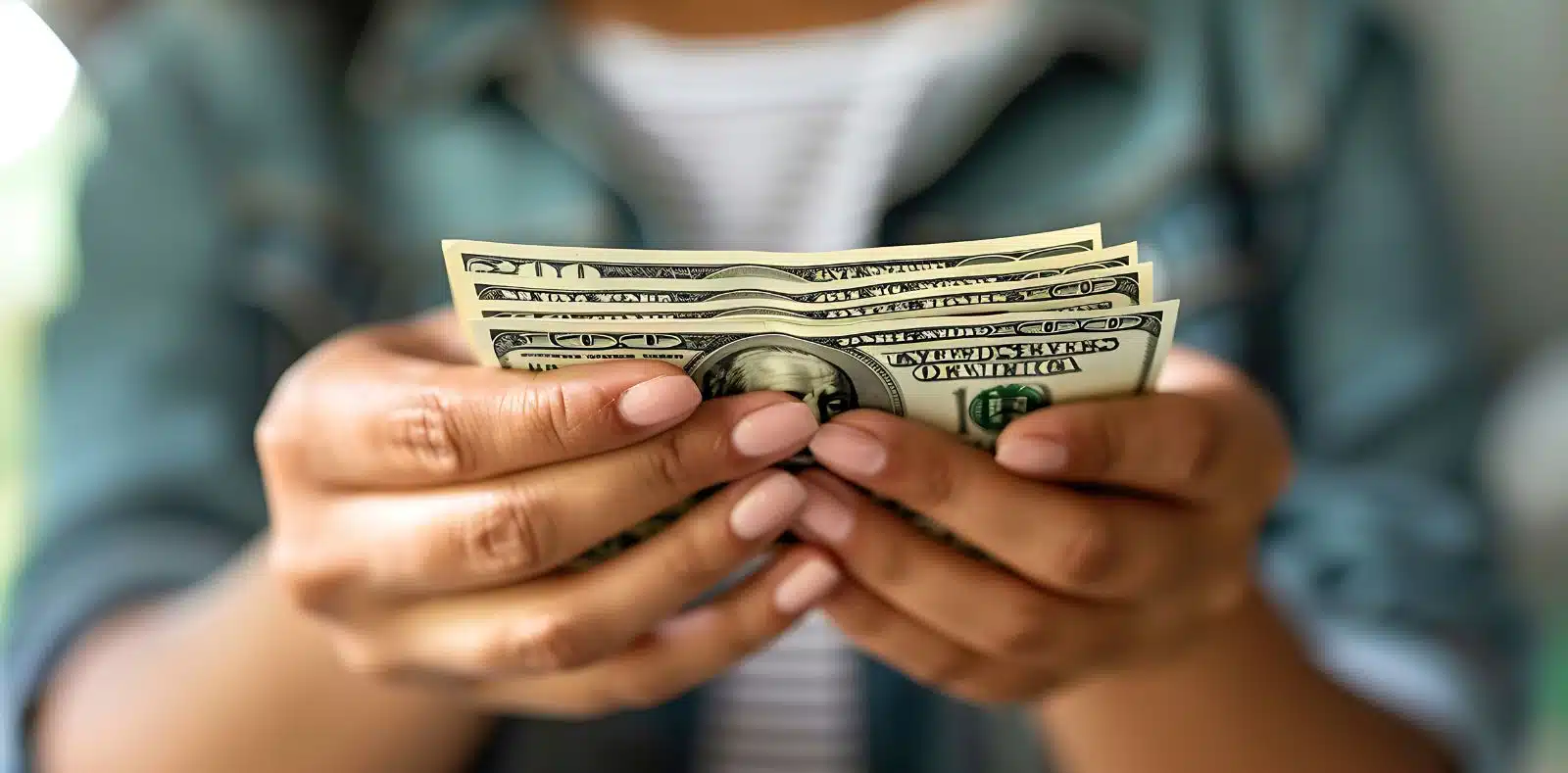 A smiling young woman in a pink jumper holds a fan of $100 bills and points at them against a teal background