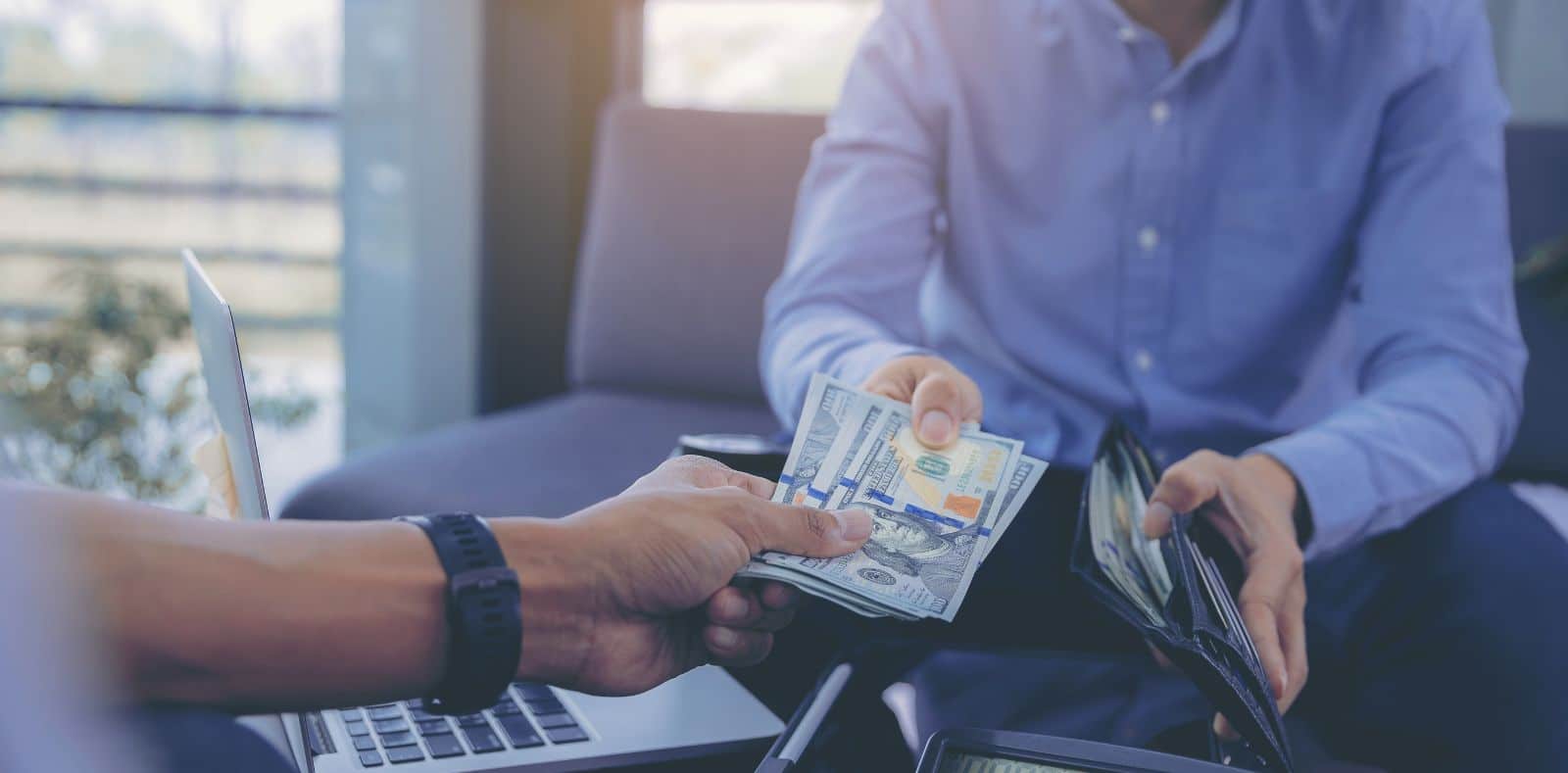A laughing bearded man in a blue suit and a smiling blonde woman sit at a desk overflowing with scattered $100 bills in a bright office