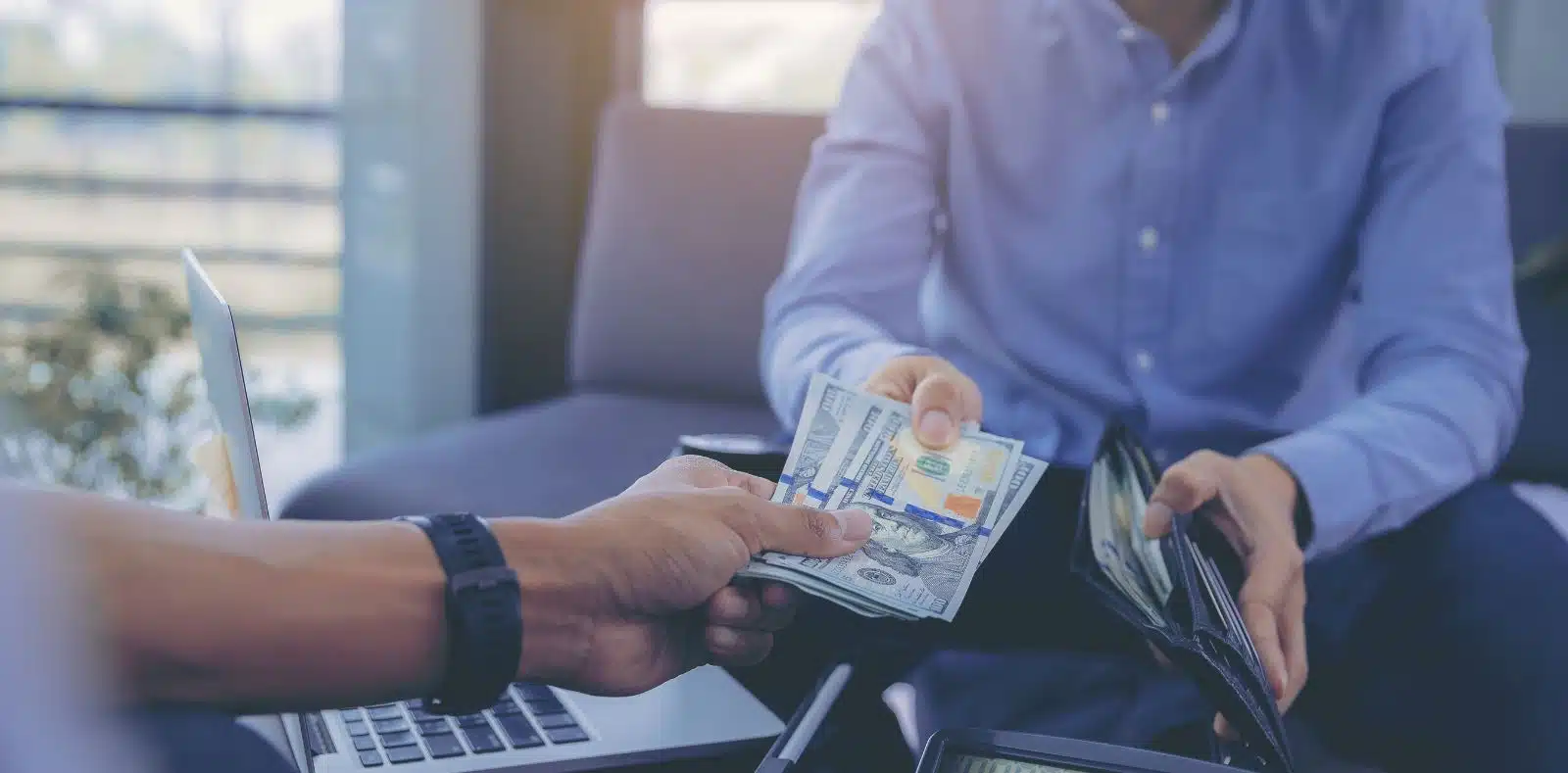 A laughing bearded man in a blue suit and a smiling blonde woman sit at a desk overflowing with scattered $100 bills in a bright office