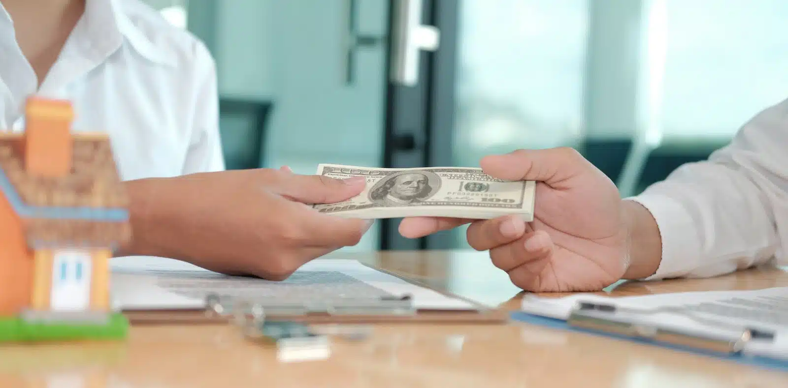 A woman in a cream cardigan and polka dot top holds several dollar bills at a desk with a laptop and papers in a sunlit workspace