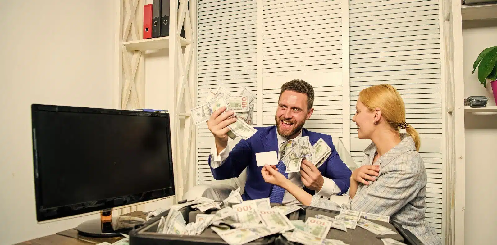 A person in a light blue shirt extends a large fan of $100 bills toward the camera with a white piggy bank and scattered money on a dark desk