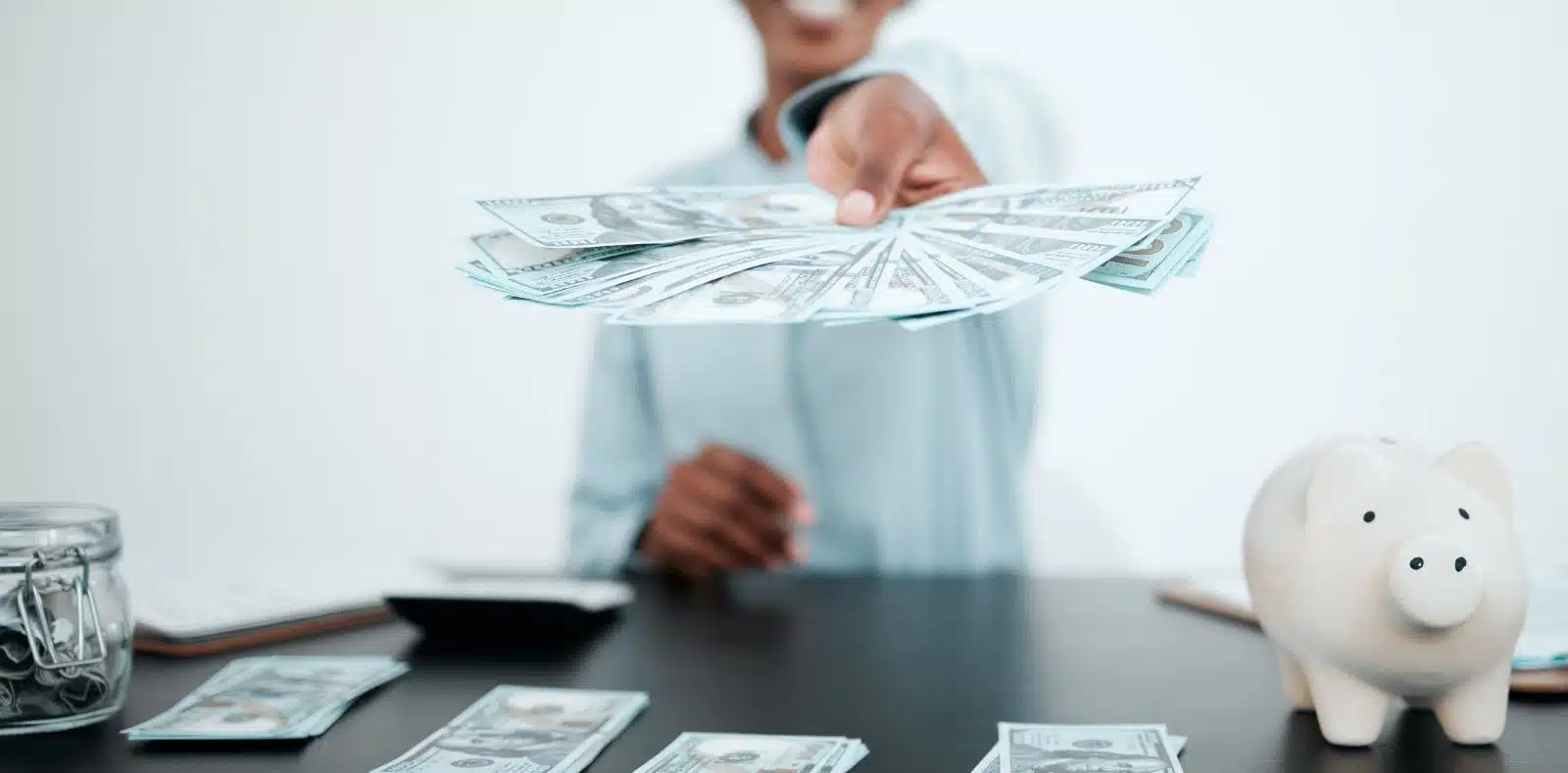 A man in a blue shirt pulls dollar bills from a leather wallet to hand to another person near a laptop in a modern office setting