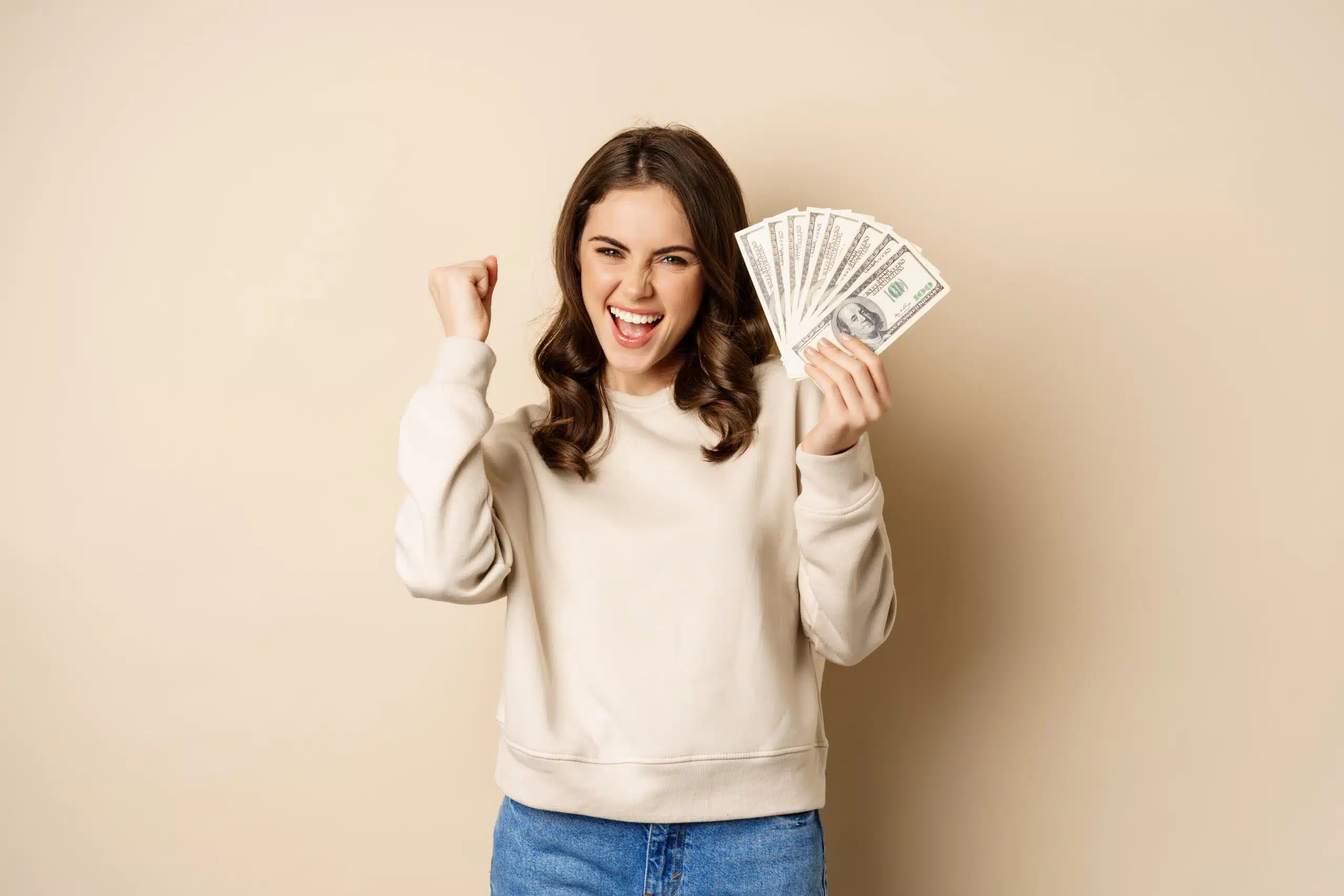 Smiling woman celebrating while holding several bills in her hand against a neutral background.